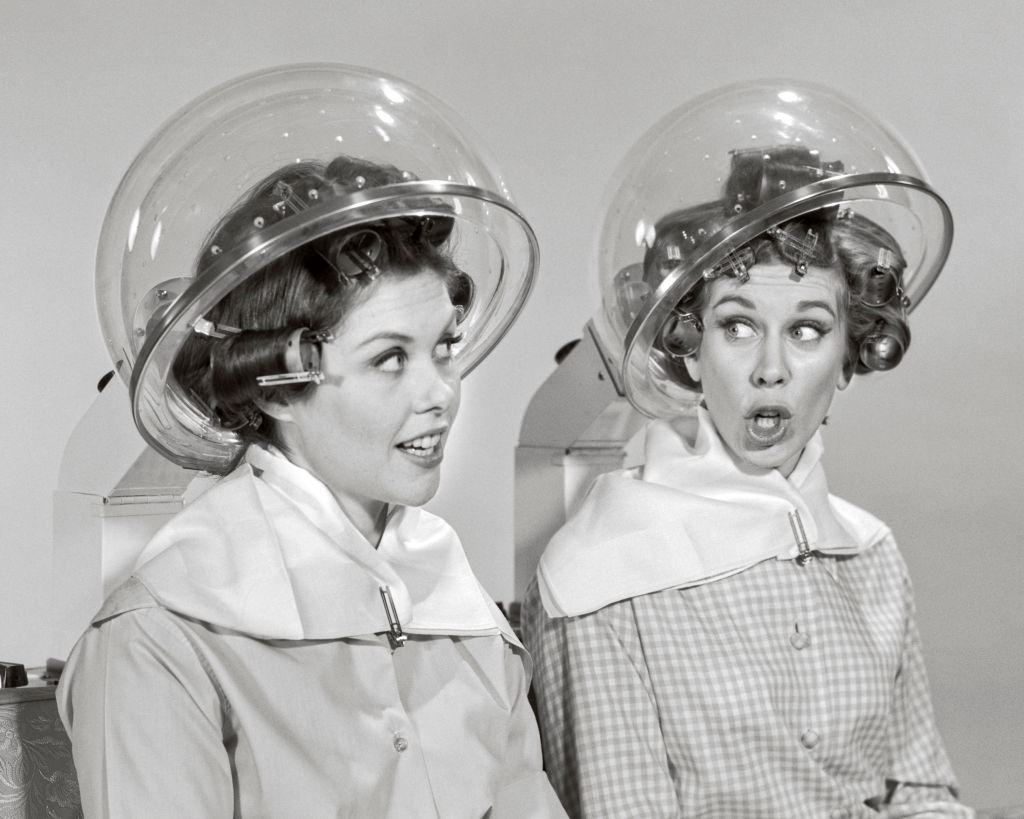 Two women rollers curler in their hair sitting under hair salon dryers talking gossiping, 1960s