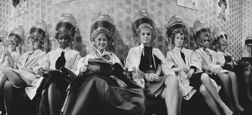 Contestants for the upcoming Miss World beauty pageant seated under hairdryers at a hairdressing salon in London on 12th November 1964.