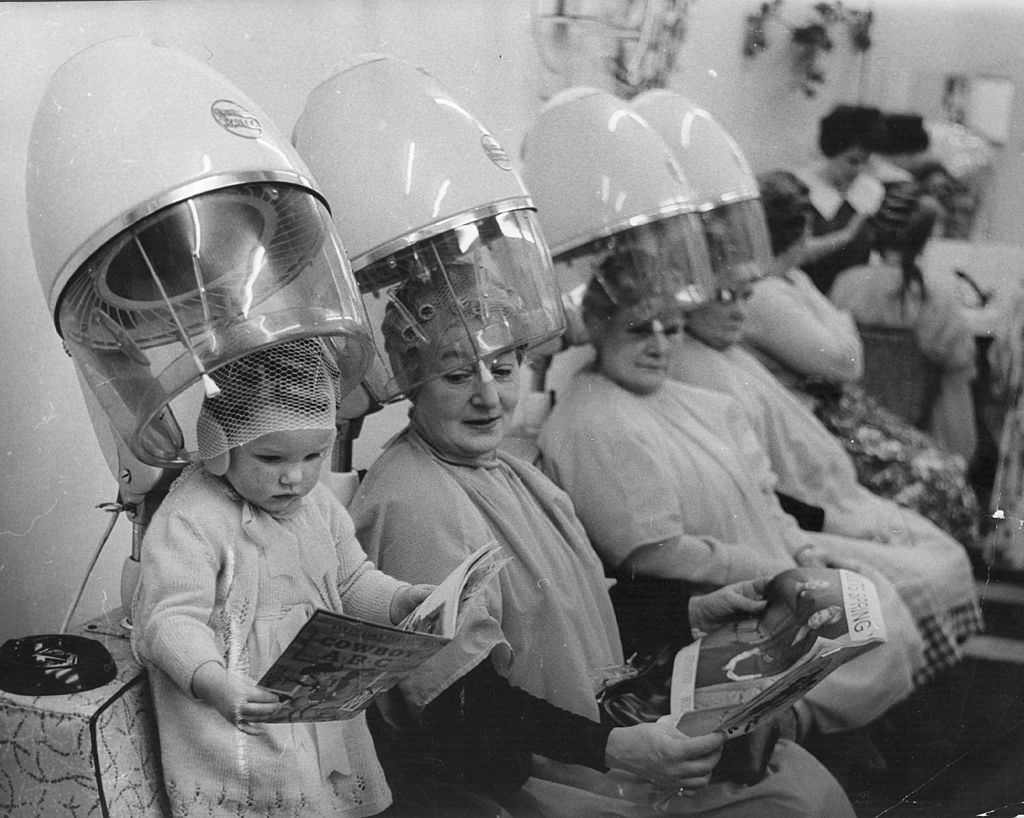 A child joins the older ladies under the hairdryers at the hairdressers.