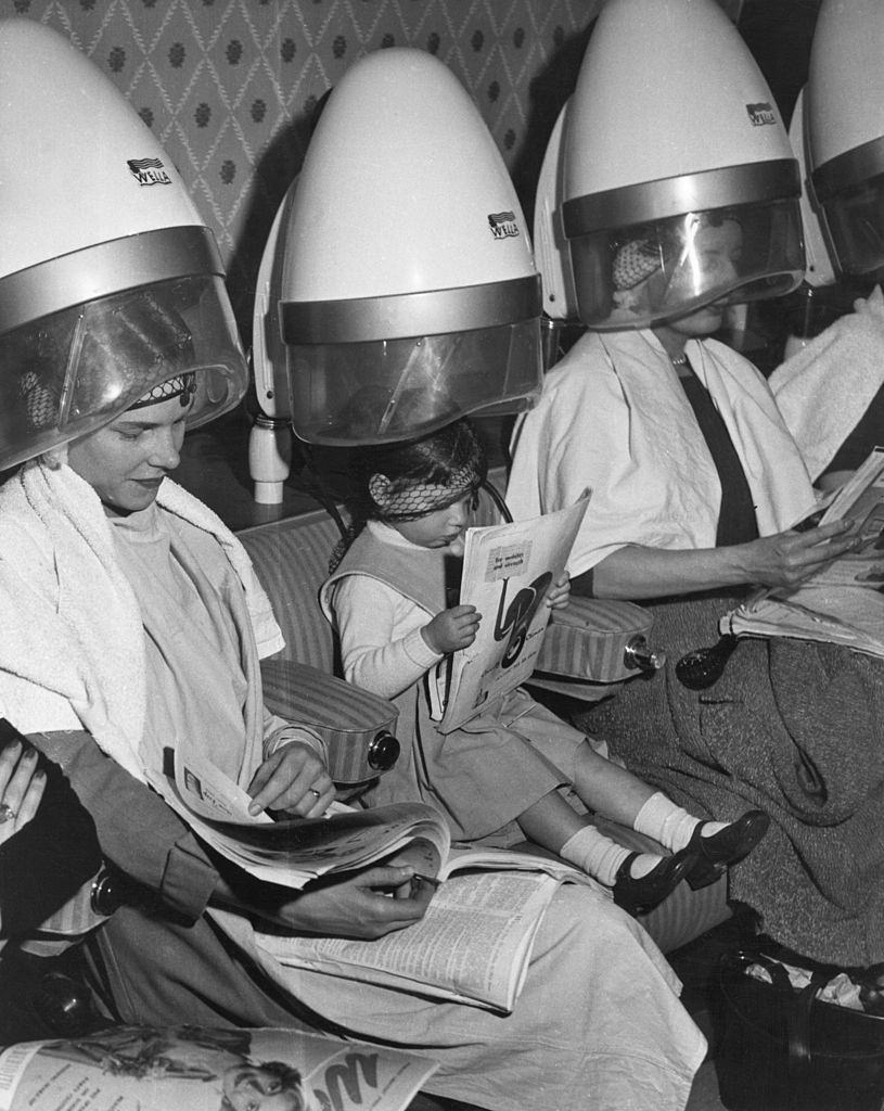 Three-year-old Jaqueline Power of Maida Vale sits under the hairdryer between two older customers at a Golders Green hairdressers, 1957