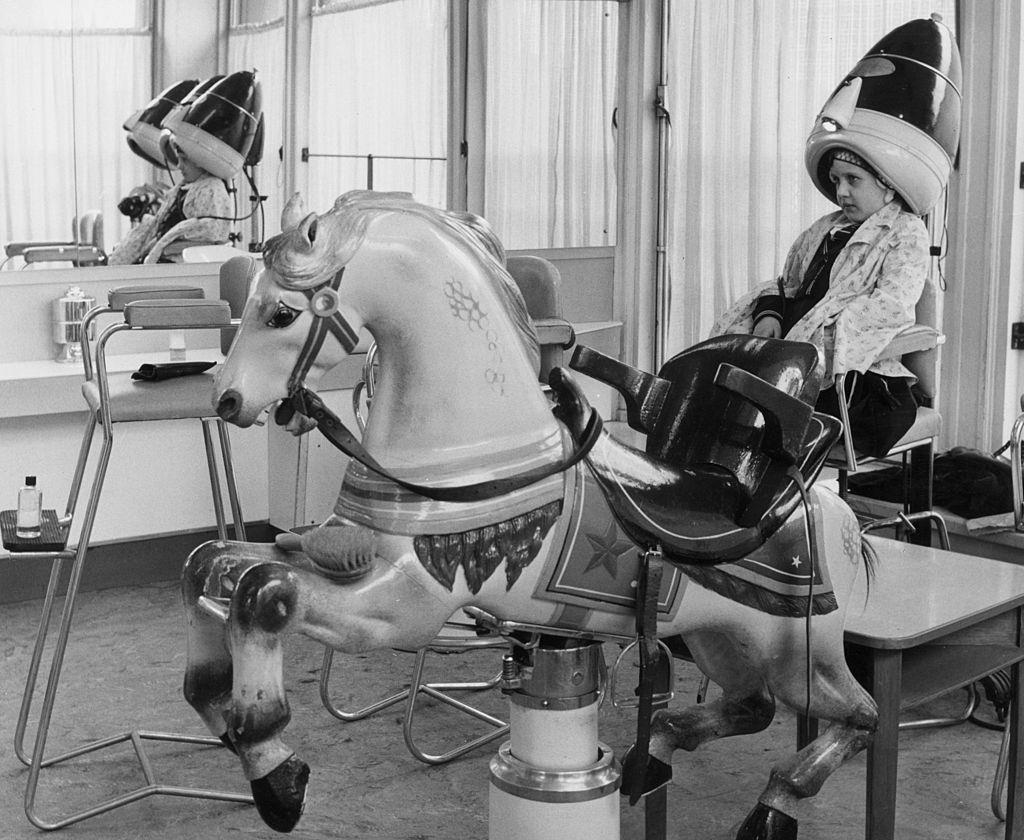 A solemn little girl sits under a hairdryer at the hairdressers which is equipped with a rocking horse for customer's amusemnt, 1957