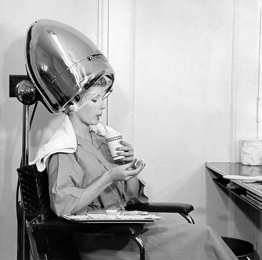 A woman drinking something under a hair drier, 1960s