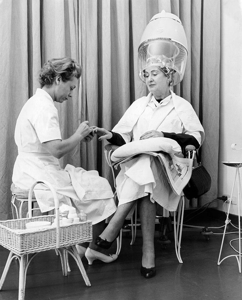 A woman with curlers under the hair dryer having a manicure, 1960s