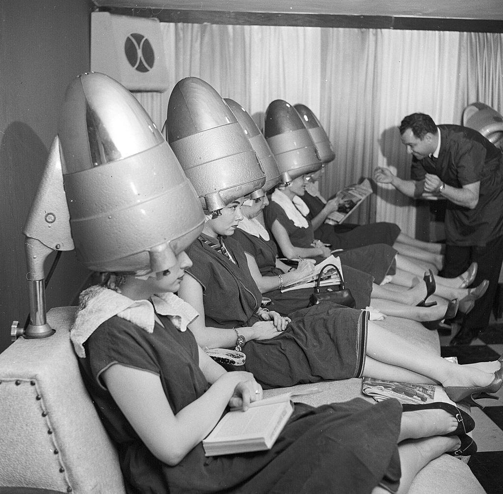 A line of women sitting under professional hairdryers in an American hairstyling salon, 1955
