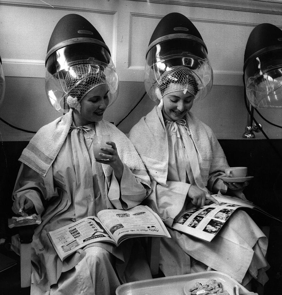 Two women sitting under hairdryers as they wait for their 'perms' to set, 1954