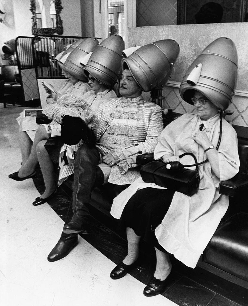 A famous television hairdresser Raymond Bessone, known as 'Teazy Weazy', sitting under a hairdryer with a group of ladies at his salon, 1969