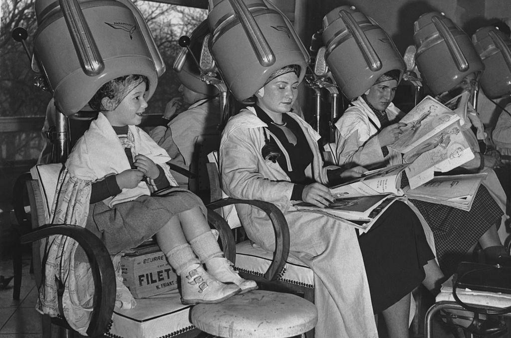 A young girl sitting on a box under a hairdryer at the salon in the 1950's.