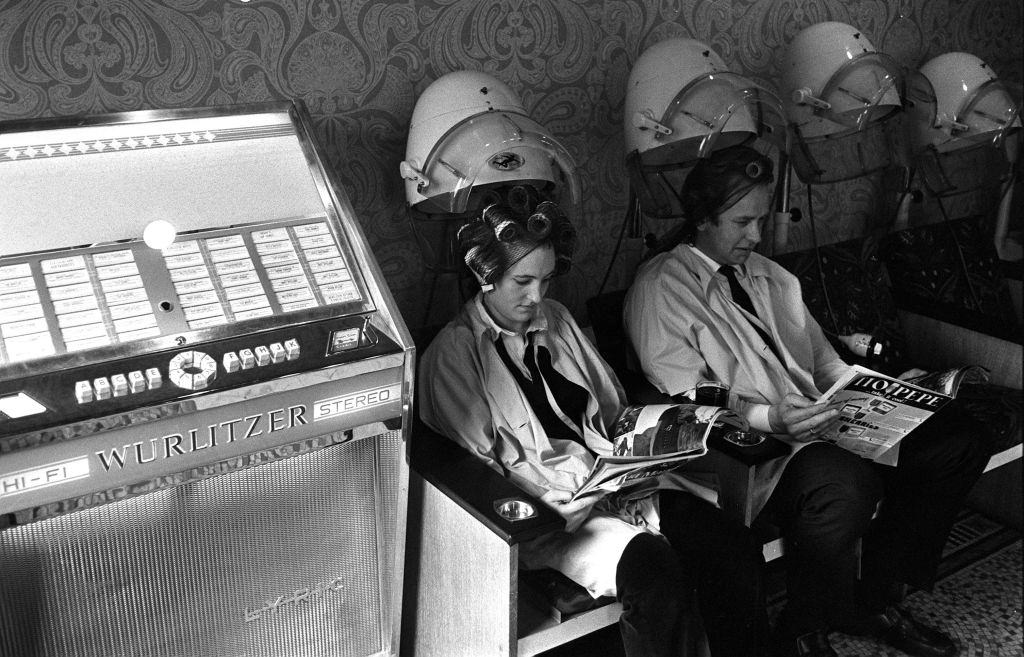 A man and woman read magazines as they sit under hairdryers at the first unisex hairdressing salon in London, as music plays from a Wurlitzer jukebox, 1970