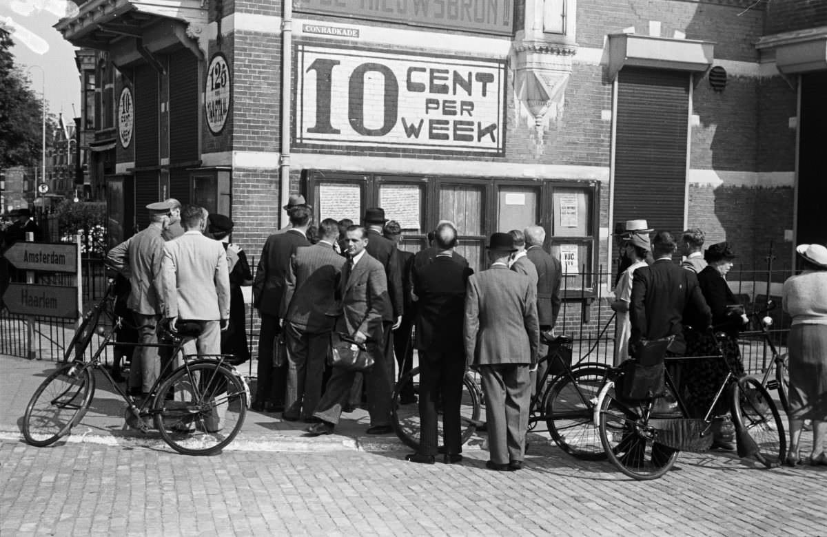 A group of people read a series of handwritten wall papers in the street, under an advertisement for the newspaper “De Nieuwsbron”.