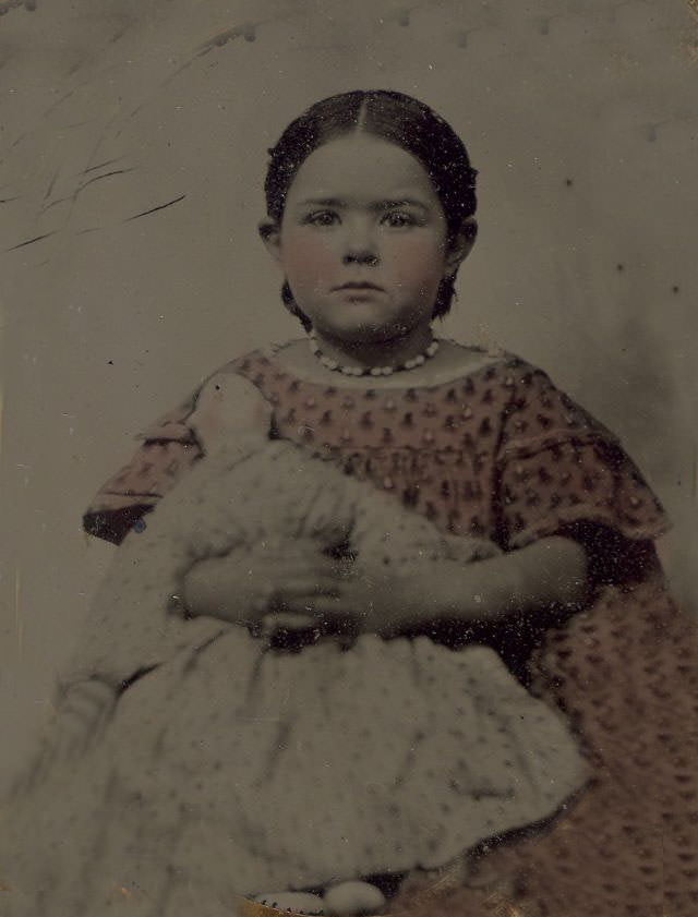 A little girl in a pink dress holding a doll. Back of image has photographer card from C.B. Lawry, Farmington, Maine