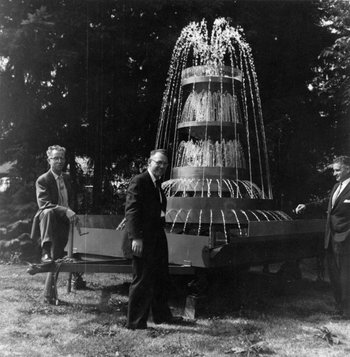 Three men gathered around a fountain, which is sitting on a trailer, 1960s