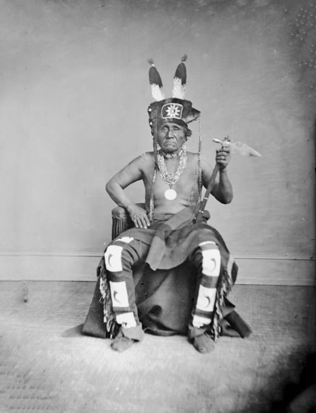 Tcha-Wan-Na-Ga-He (Buffalo Chief) in Native Dress wearing fur and feather headdress and peace medal, holding pipe-tomahawk.