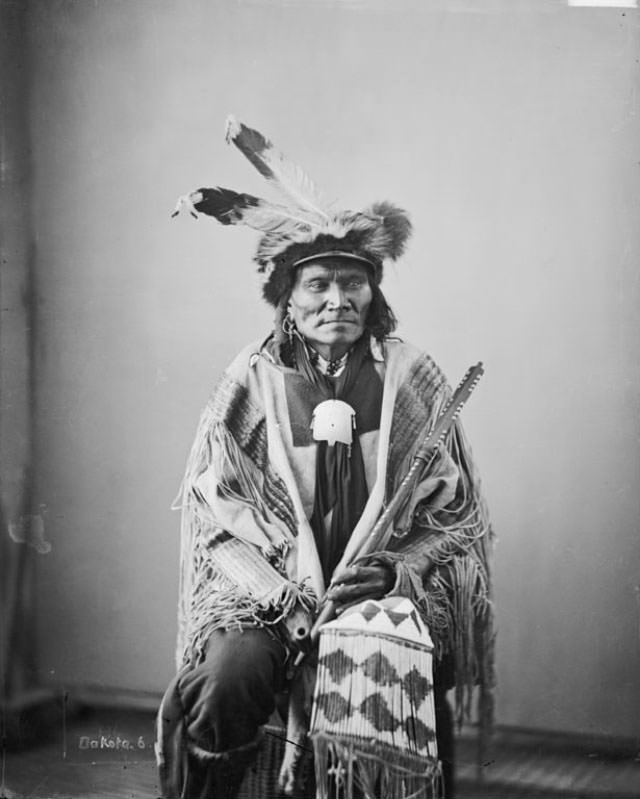 Ta-Tan-Ka-Han-Ska (Long Fox or Long Buffalo Bull) wearing skunk hat and holding pipe and beaded and quilled bag.