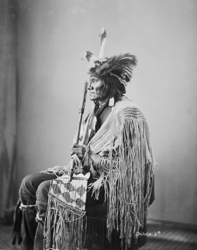Ta-Tan-Ka-Han-Ska (Long Fox or Long Buffal O Bull) in wearing feathered skunk hat and holding pipe and beaded and quilled bag.