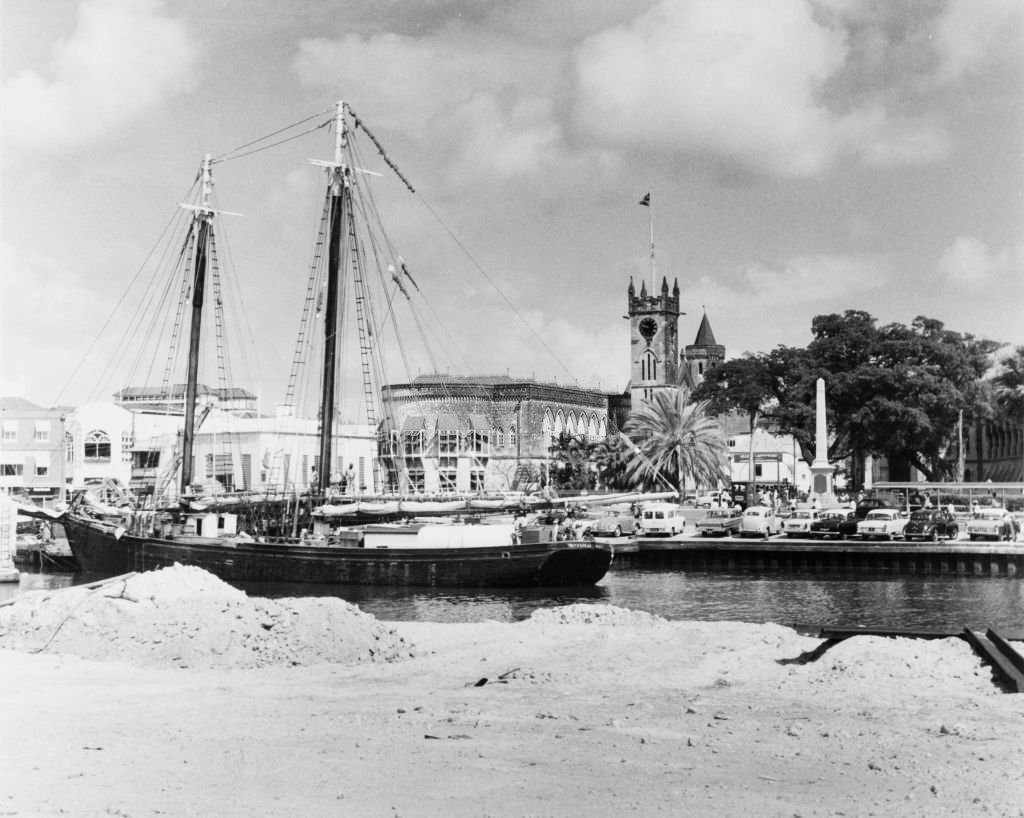 A harbour police officer talks to a man unloading bricks from a sail boat onto a truck as he patrols the Careenage. Bridgetown, Barbados, 1965.