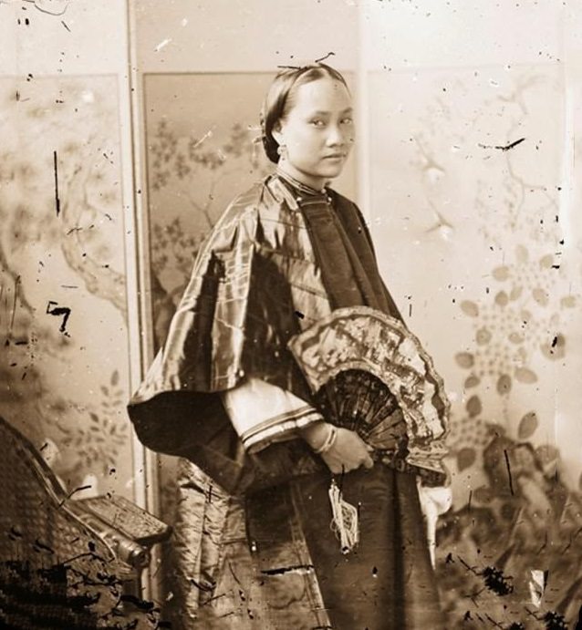 A Cantonese beauty holds an open painted fan and stands before a folding screen Guangdong, 1869