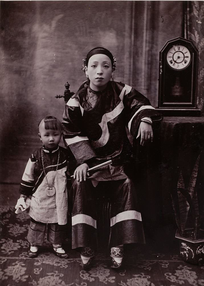A woman with bound feet sits on a chair while her small son stands next to her.