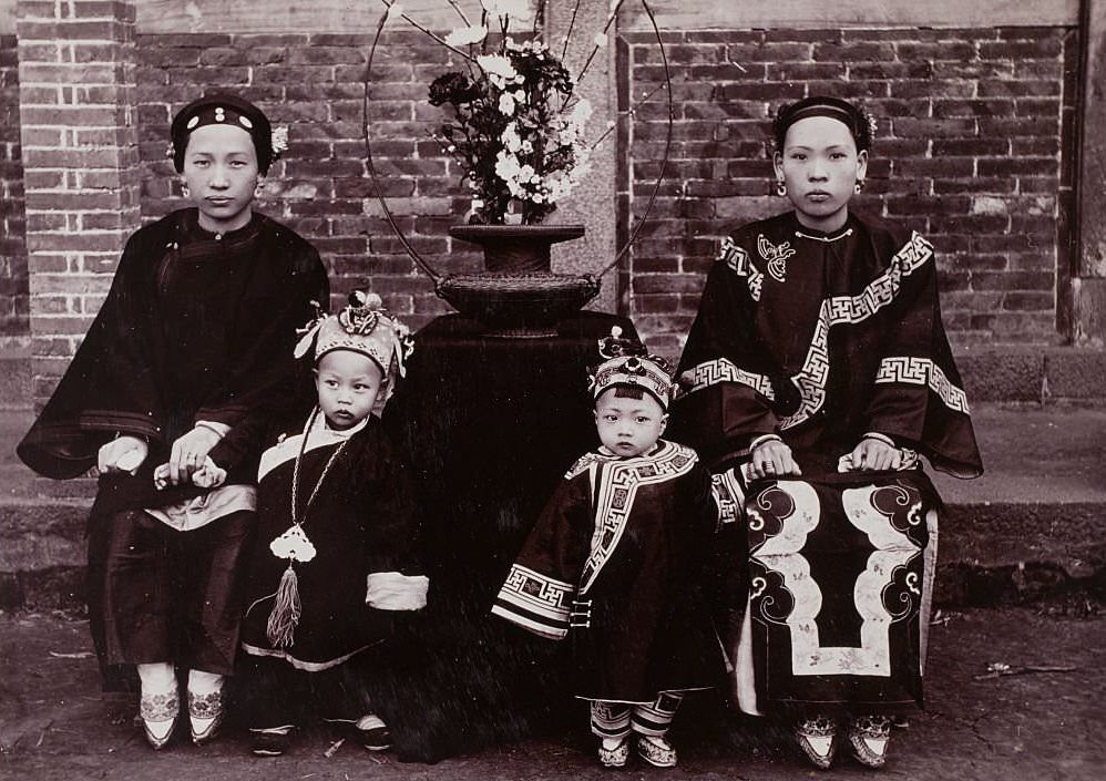 Two Chinese women with bound feet sit in chairs while their young sons, dressed in robes and hats, stand next to them.