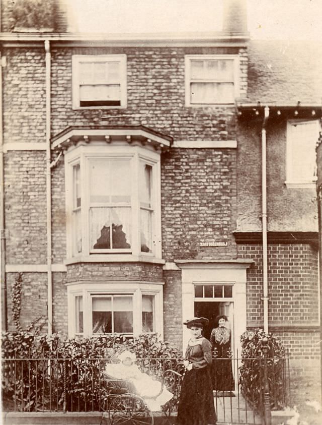 An Edwardian lady with a baby in a pram stand on the pavement outside