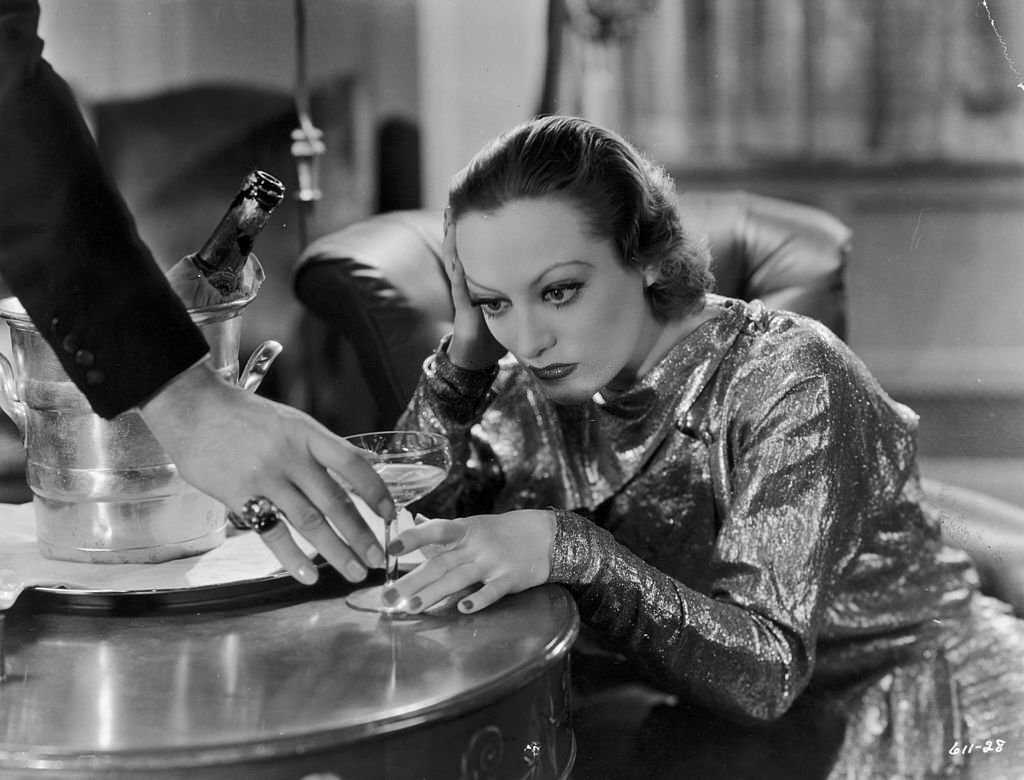 Joan Crawford sits with her head in her hand as a man reaches to take her champagne glass, 'Grand Hotel'.