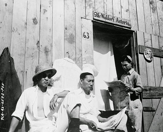 Three interned Japanese men sitting by a wooden building with the words "The Waldorf Astoria" written over the doorway.