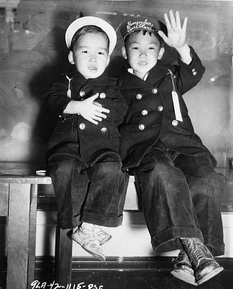 Evacuation - Two Japanese boys, one with strip "Remember Pearl Harbor" on his hat, wave good-bye [while] awaiting the bus.