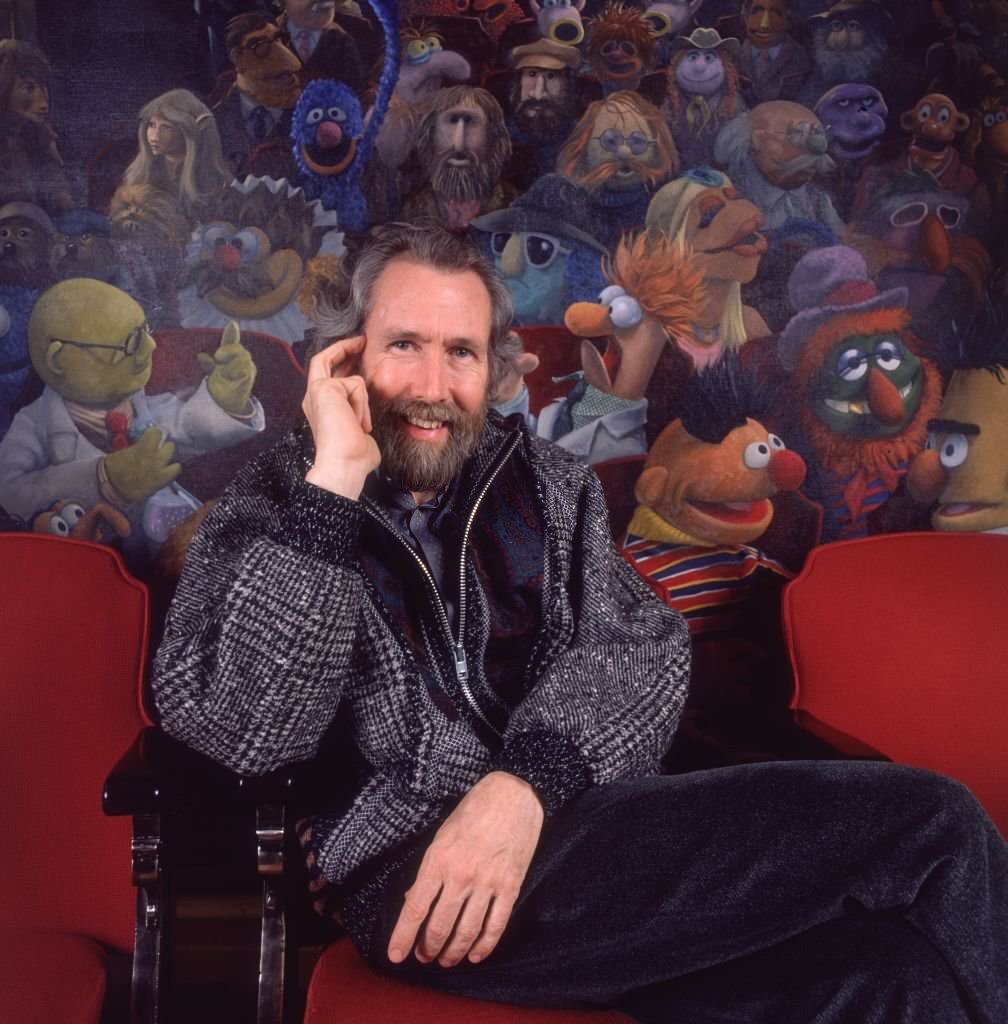 Jim Henson sitting in a theater chair in front of a painting of his creation, The Muppets.