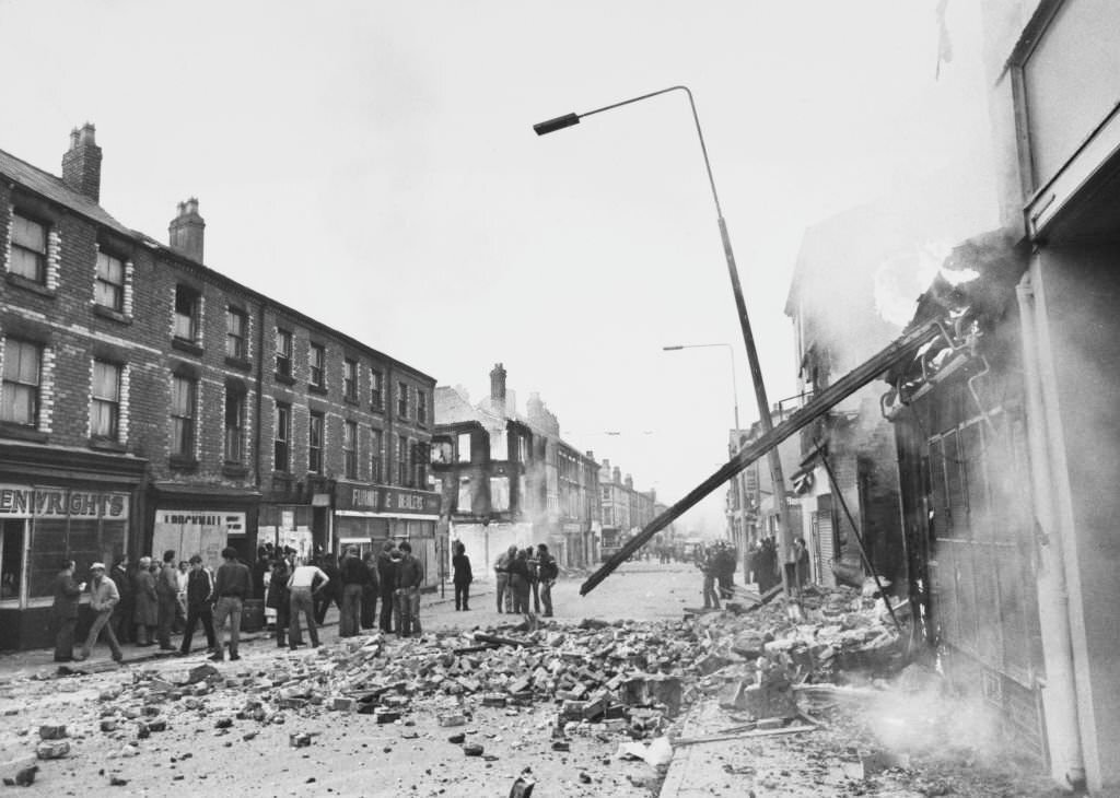 A crowd in the street stand in a brick-strewn road, beside a damaged shop, smoke pouring from the damaged facade, and a leaning lamppost in the Toxeth area of Liverpool, England, July 1981.