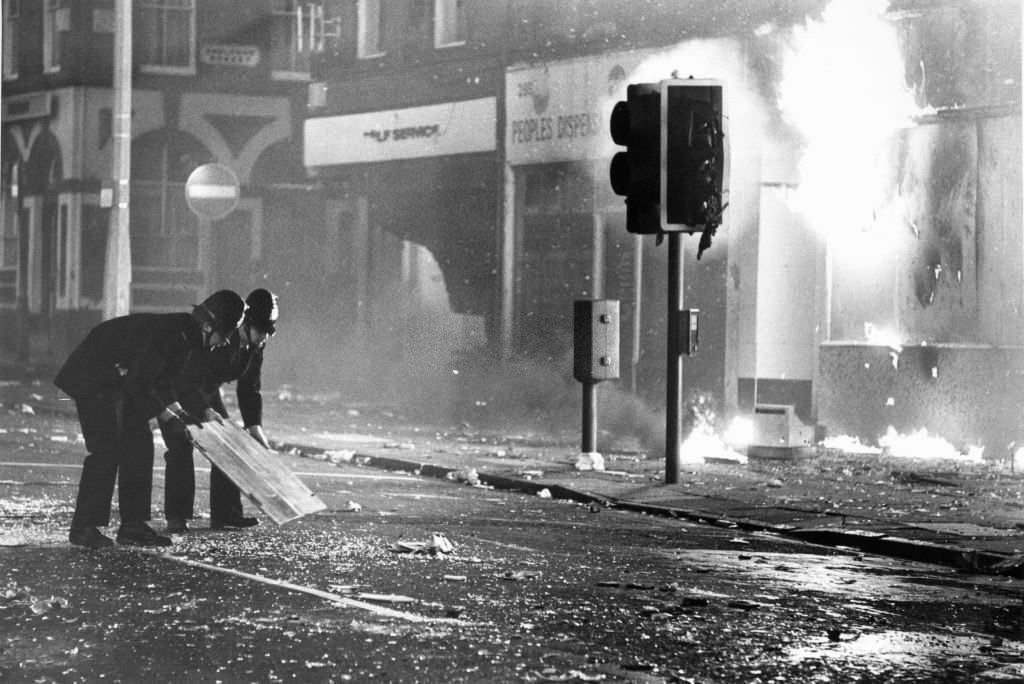 Police officers try to clear broken glass from Park Road outside a burning looted shop, so that the fire brigade can control the fire.