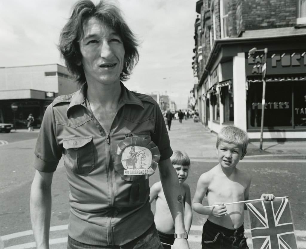 A man wearing a rosette with the message 'No Surrender' accompanied by two young boys carrying a union jack flag on the streets of Liverpool, June 1982.