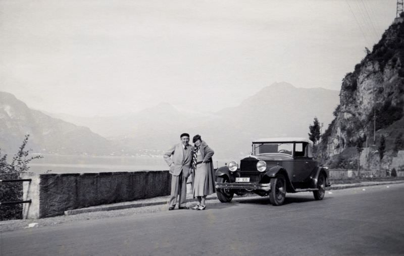 An elderly couple posing with a 1930 Packard Standard Eight Convertible Coupe on a lakeside road in autumntime.