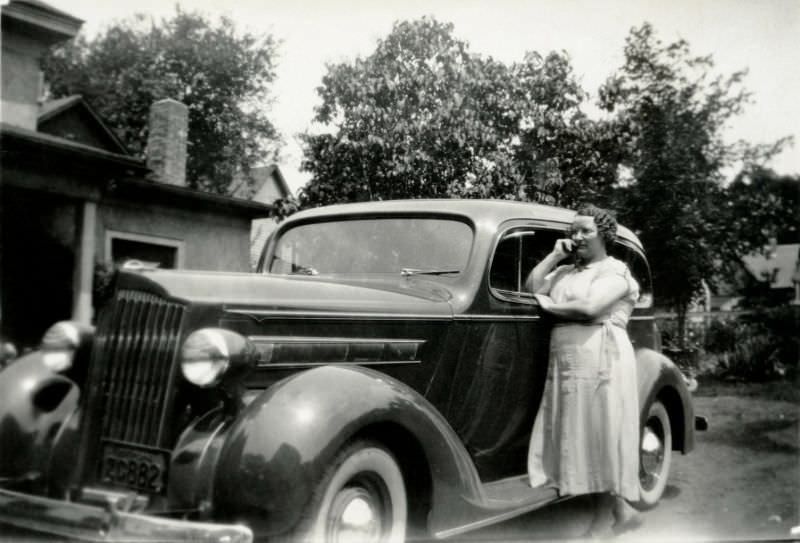 A curly-haired lady in a white linen dress posing with a 1936 Packard 120 Sedan in front of the family home, 1938