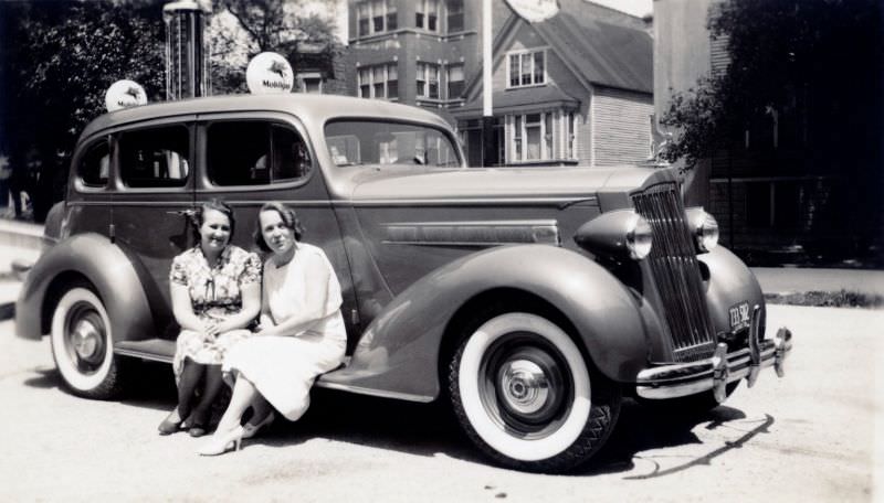 Two cheerful young ladies posing on the running board of a 1936 Packard 120 Touring Sedan at a Mobile filling station in summertime.