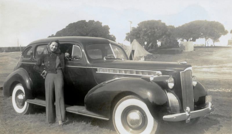 A cheerful young lady in pants with a cigarette in her left hand posing with a 1940 Packard "120" Touring Sedan.