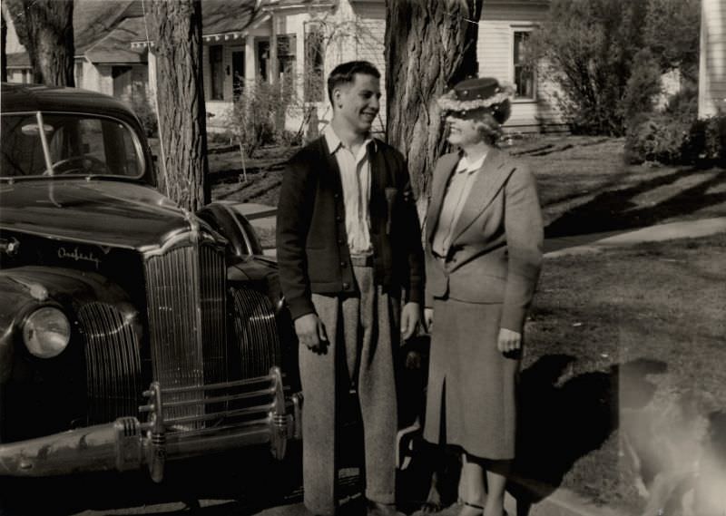 A lady wearing a female suit and a fancy hat and a man in a woolen cardigan posing in front of a 1941 Packard One Sixty, 1946