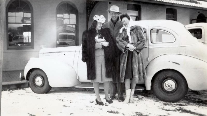 Two extravagantly dressed ladies in fur coats and a fellow with a fedora hat posing with a somewhat scruffy 1941 Packard "120" Sedan on a snowy winter's day, 1946