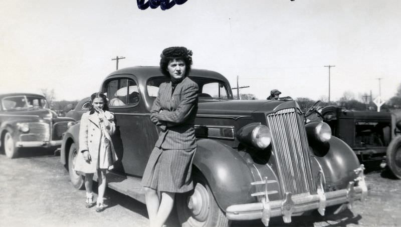 A slightly grumpy-looking lady and her daughter posing with a 1936 Packard 120 Coupe on a dirt road in the countryside.