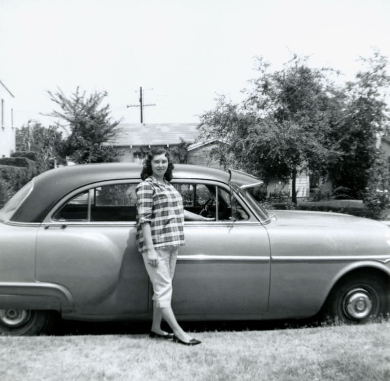 A young expectant lady dressed in Capri pants and a checkered top posing with a 1951-52 Packard 200 Sedan in a suburban street, 1955