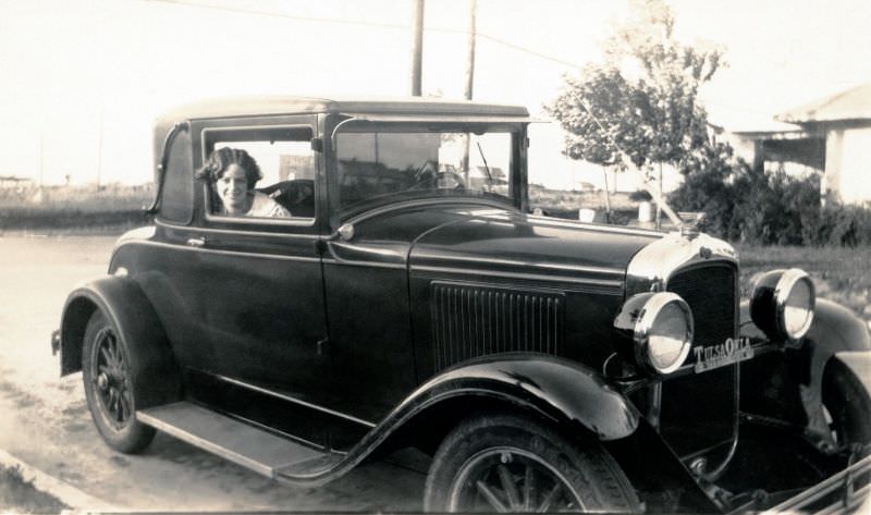A cheerful young lady posing in the passenger seat of a 1928 Pontiac Landau Coupe in a suburban street.