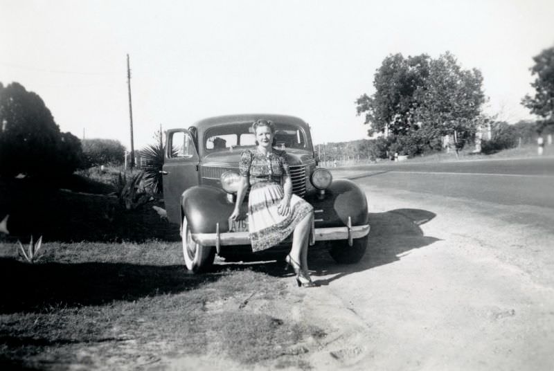 A blonde lady posing on the bumper of a 1938 Pontiac Six on the side of a country road in summertime.