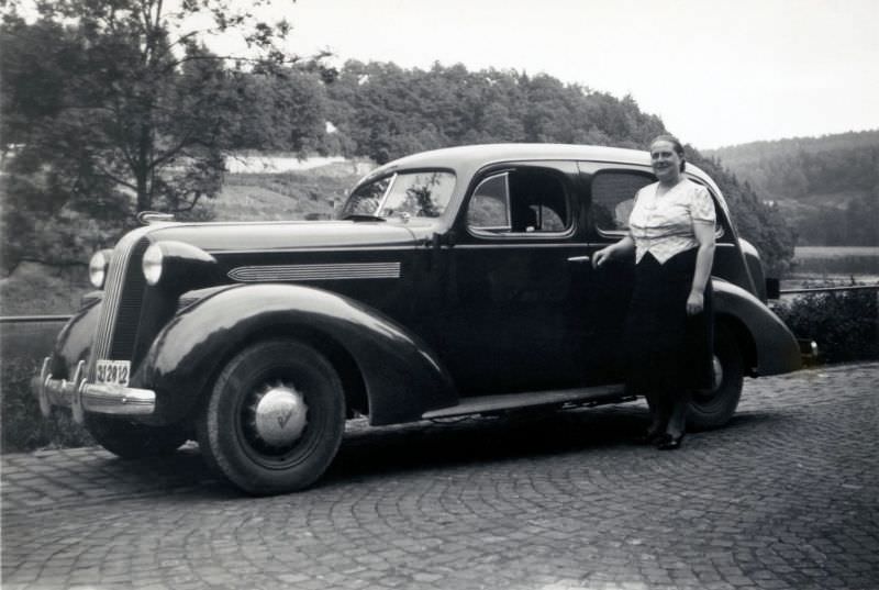 A middle-aged lady proudly posing with a 1936 Pontiac De Luxe 6 on a cobbled road in the countryside.