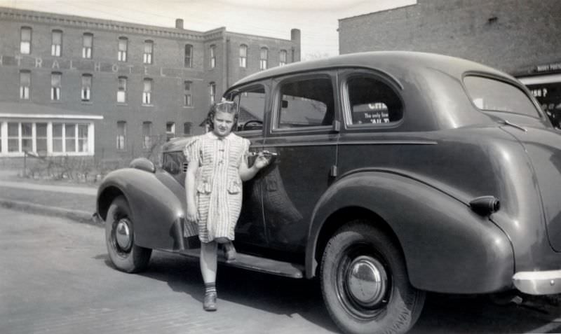 An adolescent girl in a striped dress posing on the running board of a 1939 Pontiac Sedan.