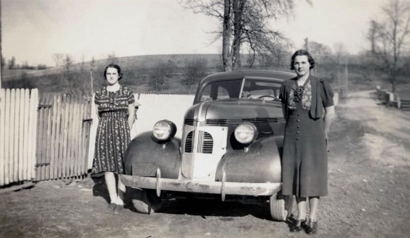 Two bespectacled ladies posing with a 1937 Pontiac on a dirt road in rural America.