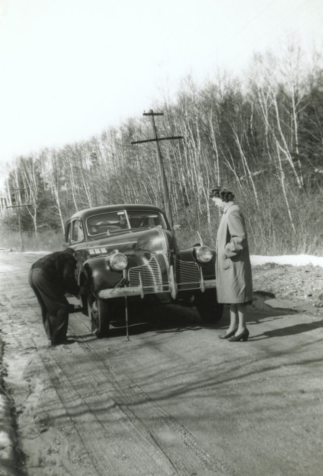 A fellow changing a tyre on a 1940 Pontiac Four-Door Sedan, while his wife (and the photographer) are looking on.