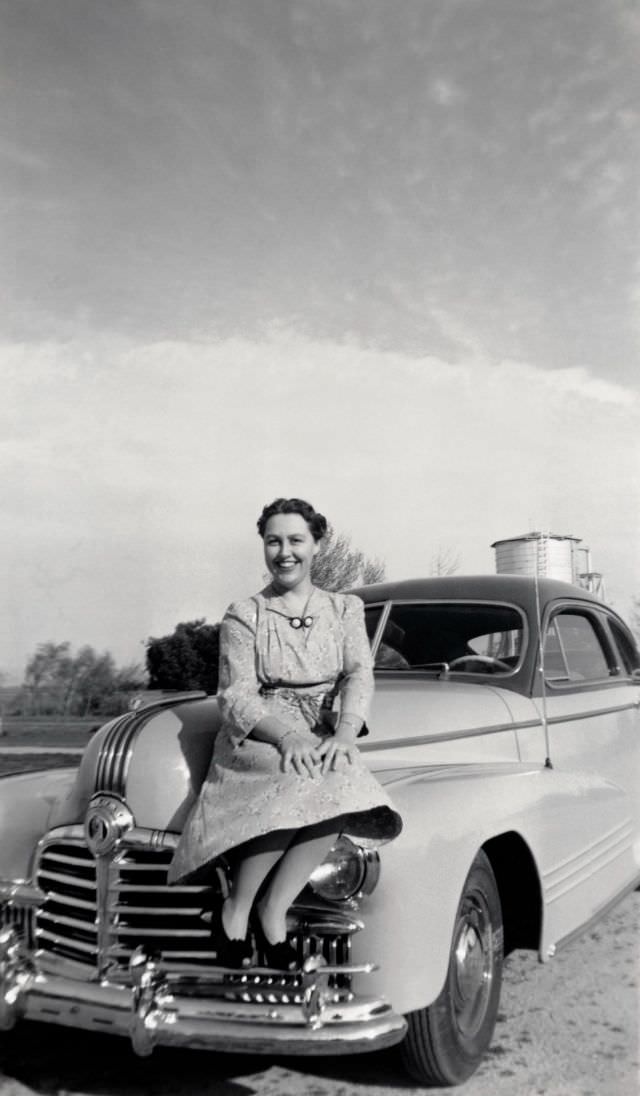 A cheerful young lady in a floral dress posing on the fender of a 1942 Pontiac Streamliner Sedan Coupe.