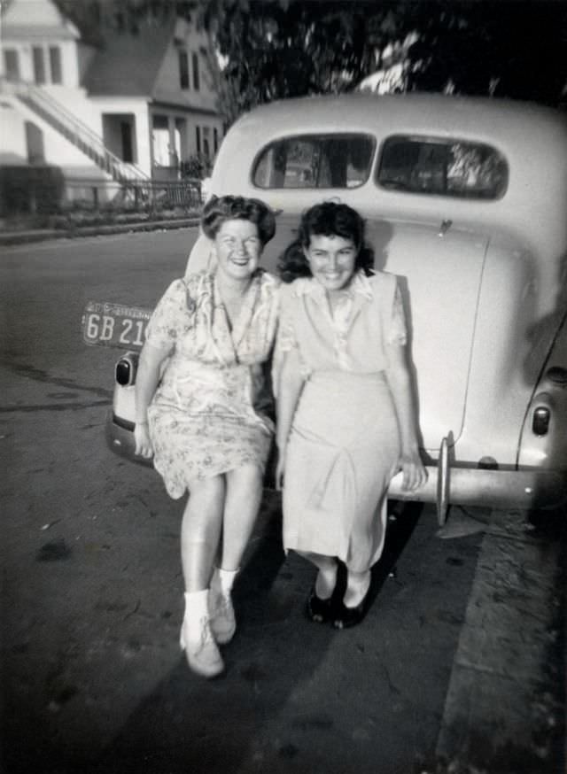 Two cheerful young ladies posing with a light-colored 1937 Pontiac in a suburban street, 1942