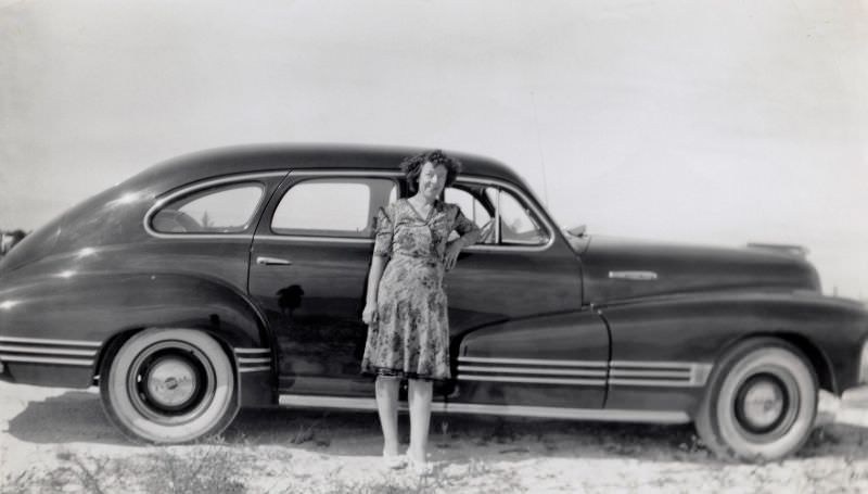 A middle-aged lady in a floral dress posing with a 1946 Pontiac Streamliner Sedan on a sunny day in the countryside, 1948