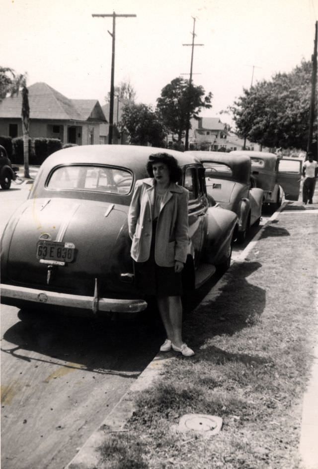 A dark-haired girl wearing white moccasins and a knee-length skirt posing with a 1940 Pontiac Special Six in a residential street.