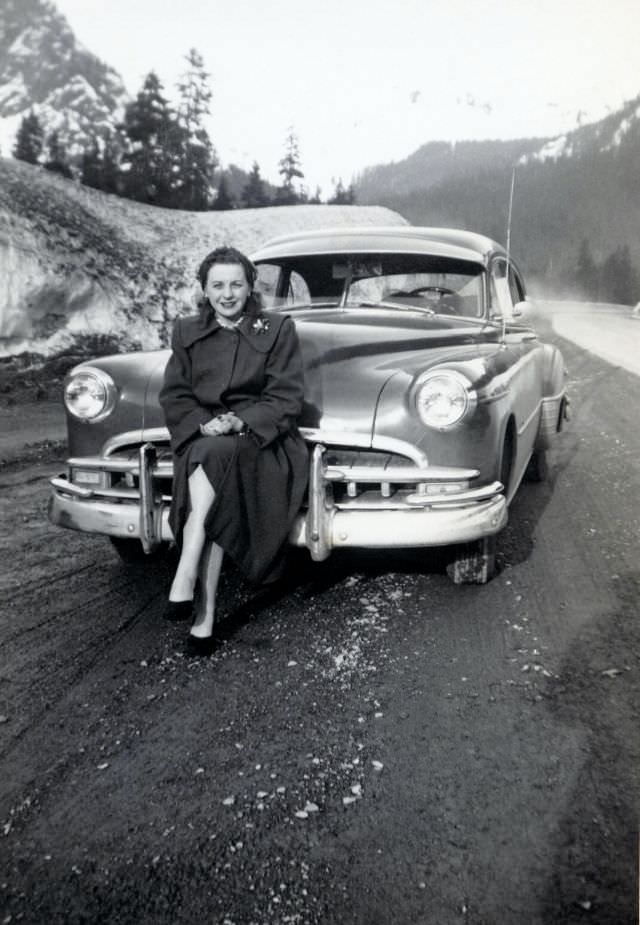 A brunette lady posing on the bumper of a 1949 Pontiac on the side of a mountain road.