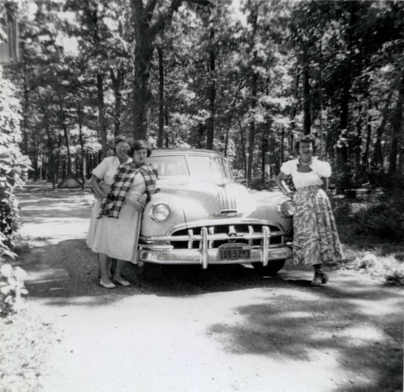 Three ladies, possibly grandmother, mother, and daughter, posing with a 1950 Pontiac Chieftain on a gravel road in a forest.